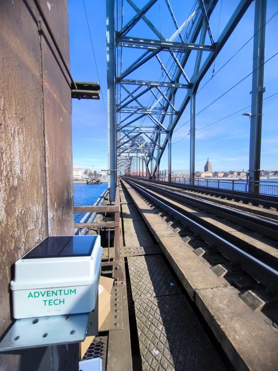Vibration wireless bridge monitoring solution Adventum Tech Vibration wireless bridge monitoring solution by Adventum Tech, featuring a solar-powered sensor mounted on a bridge structure with railway tracks, overlooking a river and cityscape under a clear blue sky.